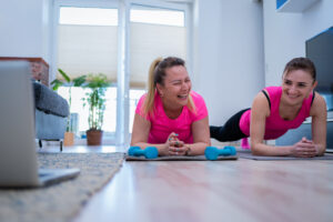 Parlak spor kıyafetleri içinde neşeli genç kadın arkadaşlar, evde mat üzerinde plank egzersizi yapmaya çalışırken dizüstü bilgisayarda video izliyor ve mutlu bir şekilde gülüyorlar